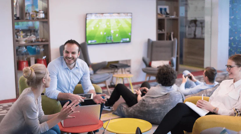 Smiling team members collaborating in a modern casual office lounge with laptops and tablets, representing engaged remote and in-person team communication in an online business setting
