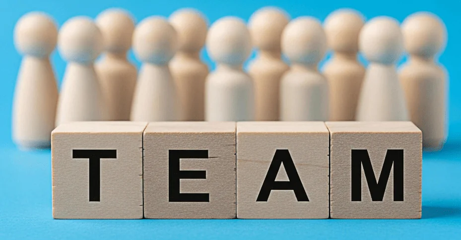 Wooden letter blocks spelling the word TEAM in front of a row of blurred wooden peg figures on a blue background, representing the concept of team unity and professional collaboration