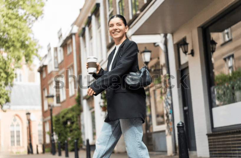 Confident woman smiling and walking with coffee on a sunny city street, representing a redefined identity grounded in personal values, truth, and authentic self-expression