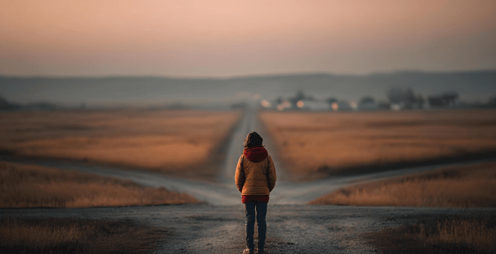 Lone person in an orange jacket standing at the end of a long empty road at sunset, representing the emotional cost of living under unquestioned labels and the longing for a new direction